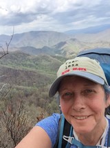 Selfie of a woman wearing a baseball cap bearing the words "Fear Less Love More", with mountains in the background. 